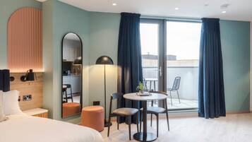 A modern hotel room with a bed, a round dining table with chairs, a small stool, a mirror, and a view of the outdoors through a sliding glass door.