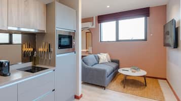 A modern kitchen with a microwave, stove, and knife block. There is a gray sofa and a small round table with books and a remote control.