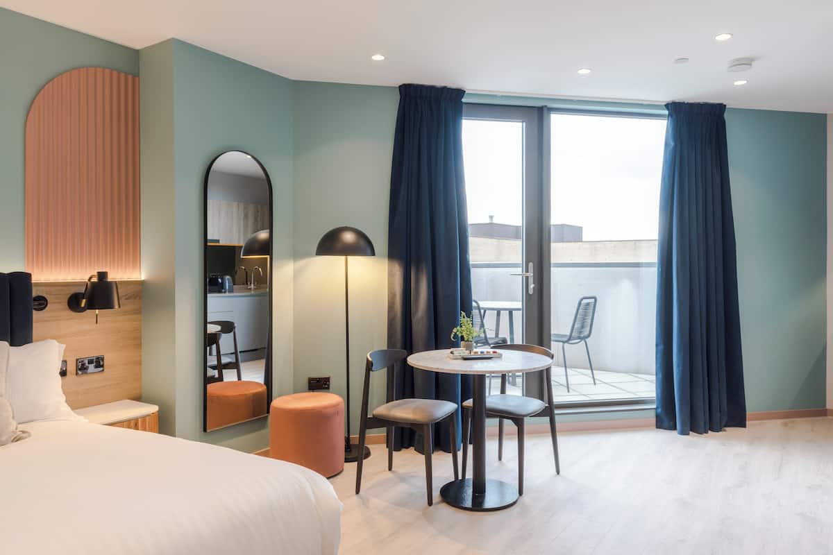 Une chambre d’hôtel moderne avec un lit, une table de salle à manger ronde entourée de chaises, un petit tabouret, un miroir et une vue sur l’extérieur grâce à une porte-fenêtre coulissante.