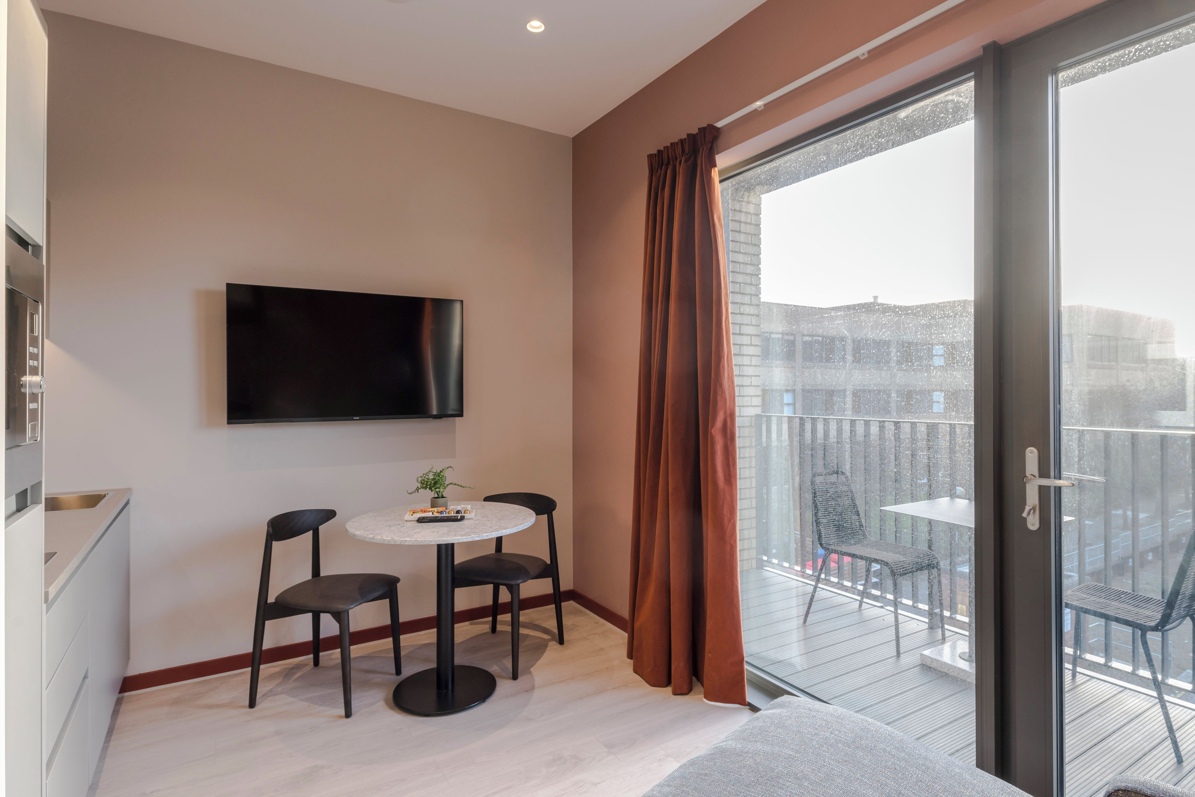 A modern room with a flat-screen TV mounted on the wall, a small round table with two chairs, and a balcony with a glass door.