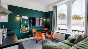 Une pièce moderne avec un mur vert, une table à manger avec des chaises, un canapé, un téléviseur et une fenêtre donnant sur des immeubles.