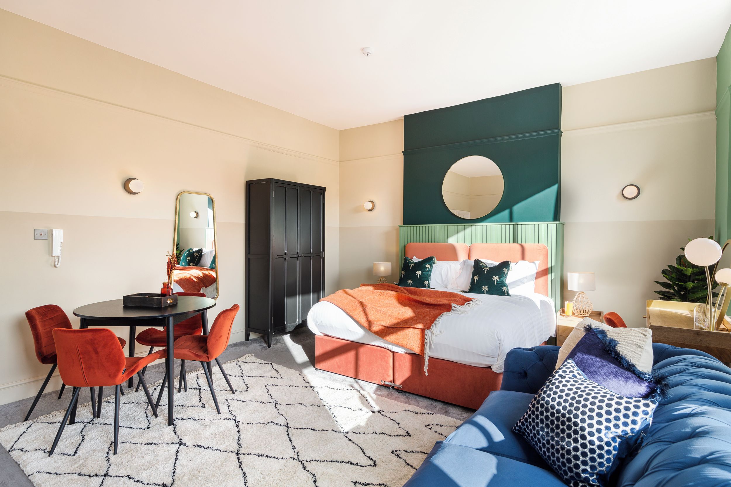 Una habitación de hotel con una cama, un espejo redondo, una mesa de comedor con sillas y un sofá azul.