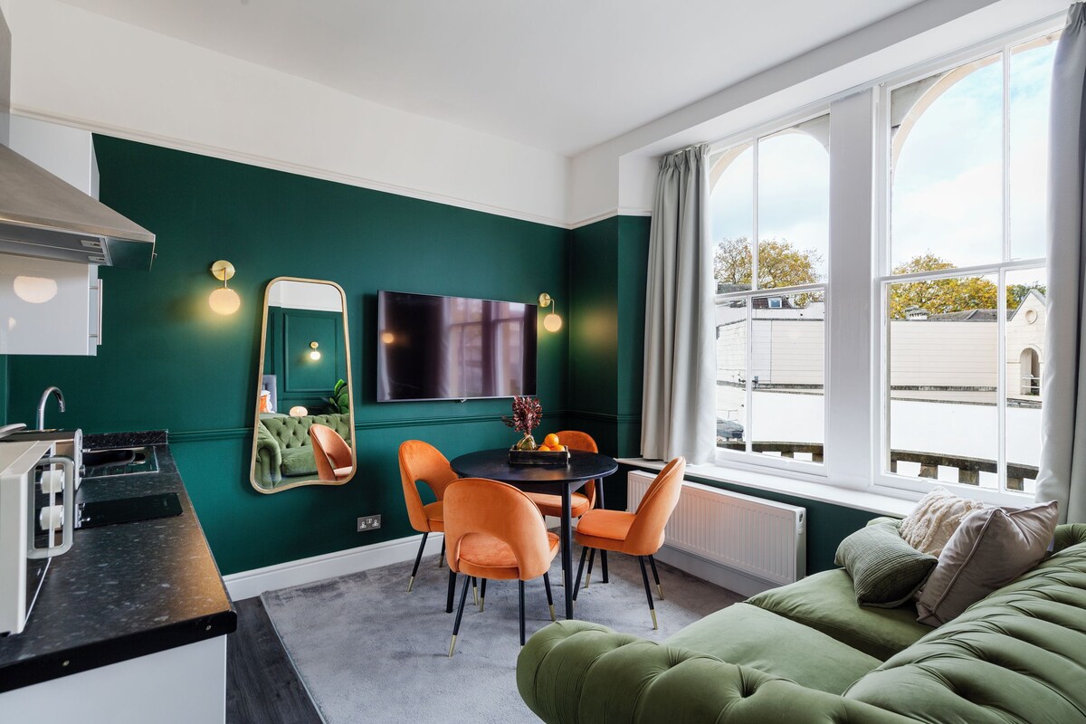 Une pièce moderne avec un mur vert, une table à manger avec des chaises, un canapé, un téléviseur et une fenêtre donnant sur des immeubles.