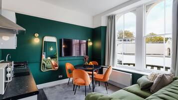 Una habitación moderna con una pared verde, una mesa de comedor con sillas, un sofá, un televisor y una ventana con vistas a edificios.