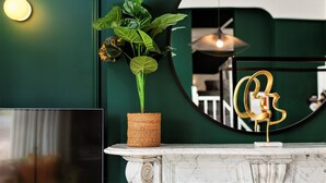 Un mur vert orné d’un miroir rond qui reflète une pièce, une plante en pot et une sculpture abstraite dorée.