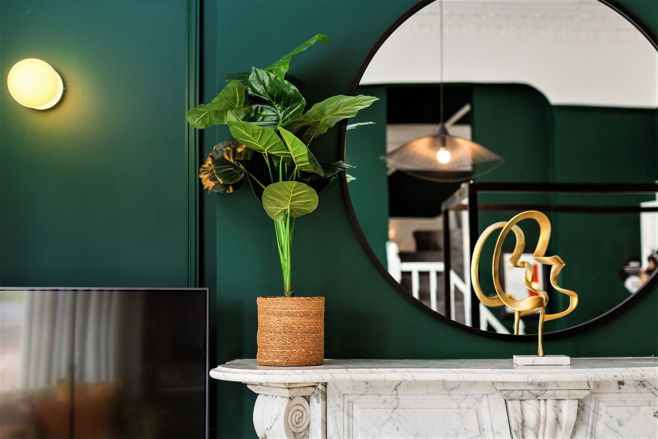 A green wall with a round mirror reflecting a room, a potted plant, and a gold abstract sculpture.