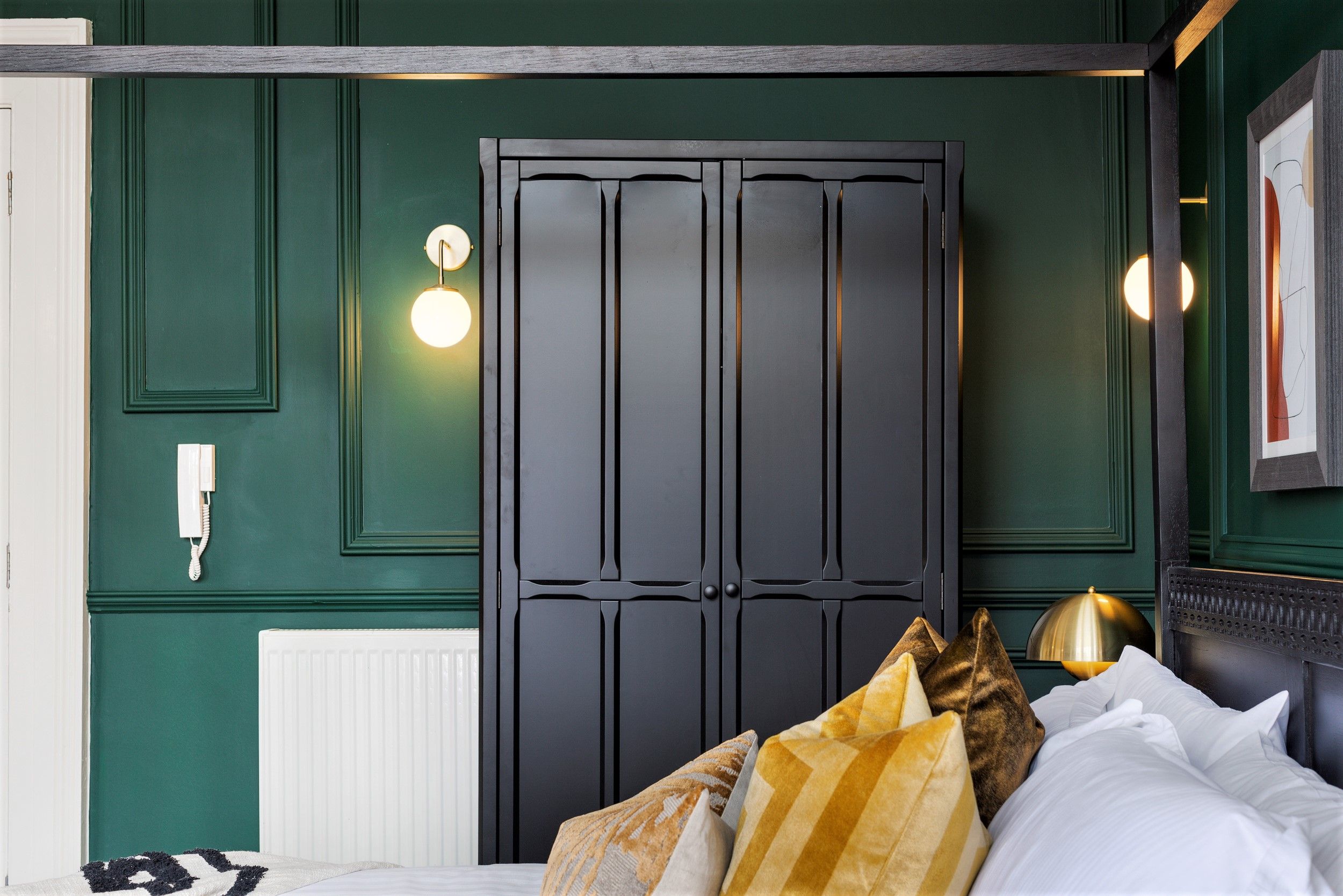A bedroom with a dark wooden wardrobe, a white radiator, a bedside table with a lamp, and a bed with multiple pillows.