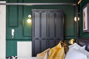 Un dormitorio con un armario de madera oscura, un radiador blanco, una mesita de noche con una lámpara y una cama con muchos almohadones.