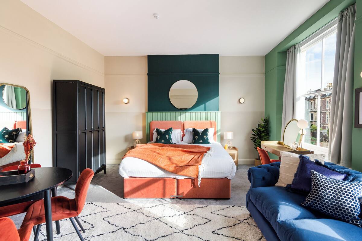 Een moderne slaapkamer met een groot bed, een blauwe bank, een eettafel en een spiegel.