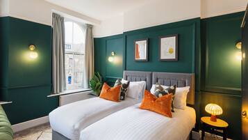 A hotel room with two beds, a window with curtains, wall-mounted lights, and framed artwork.