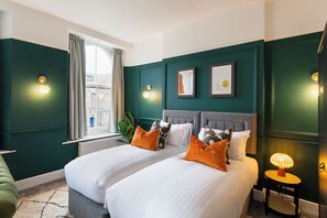 A hotel room with two beds, a window with curtains, wall-mounted lights, and framed artwork. - Clifton House - Your Apartment (Bristol)
