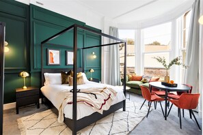 A modern bedroom with a four-poster bed, a green accent wall, and a view of the outdoors through large windows. - Clifton House - Your Apartment (Bristol)