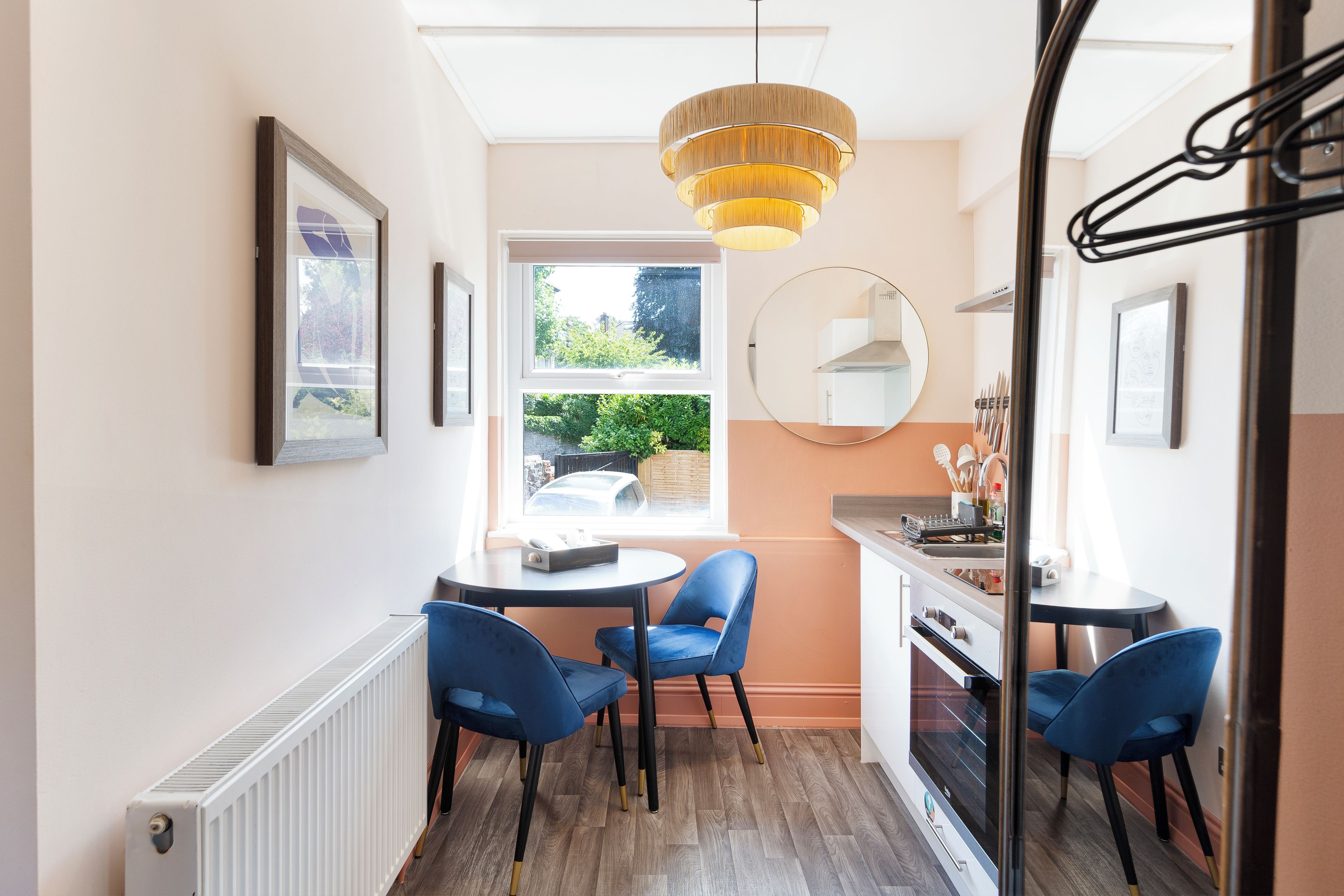 A compact kitchen with a dining area, featuring a round table, blue chairs, a hanging light fixture, and a view through the window.
