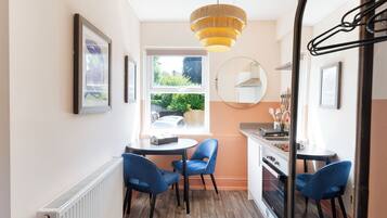 A compact kitchen with a dining area, featuring a round table, blue chairs, a hanging light fixture, and a view through the window.