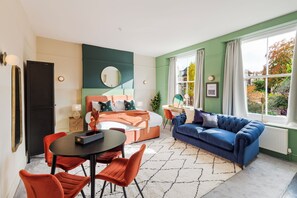 Een moderne kamer met een bed, een blauwe bank, een eettafel met stoelen en een groot raam met gordijnen.