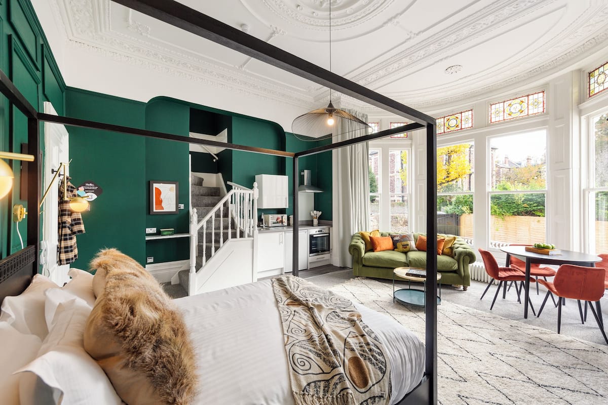 Een kamer met een hemelbed, een trap, een eethoek met een tafel en stoelen, en grote ramen met uitzicht op groen.
