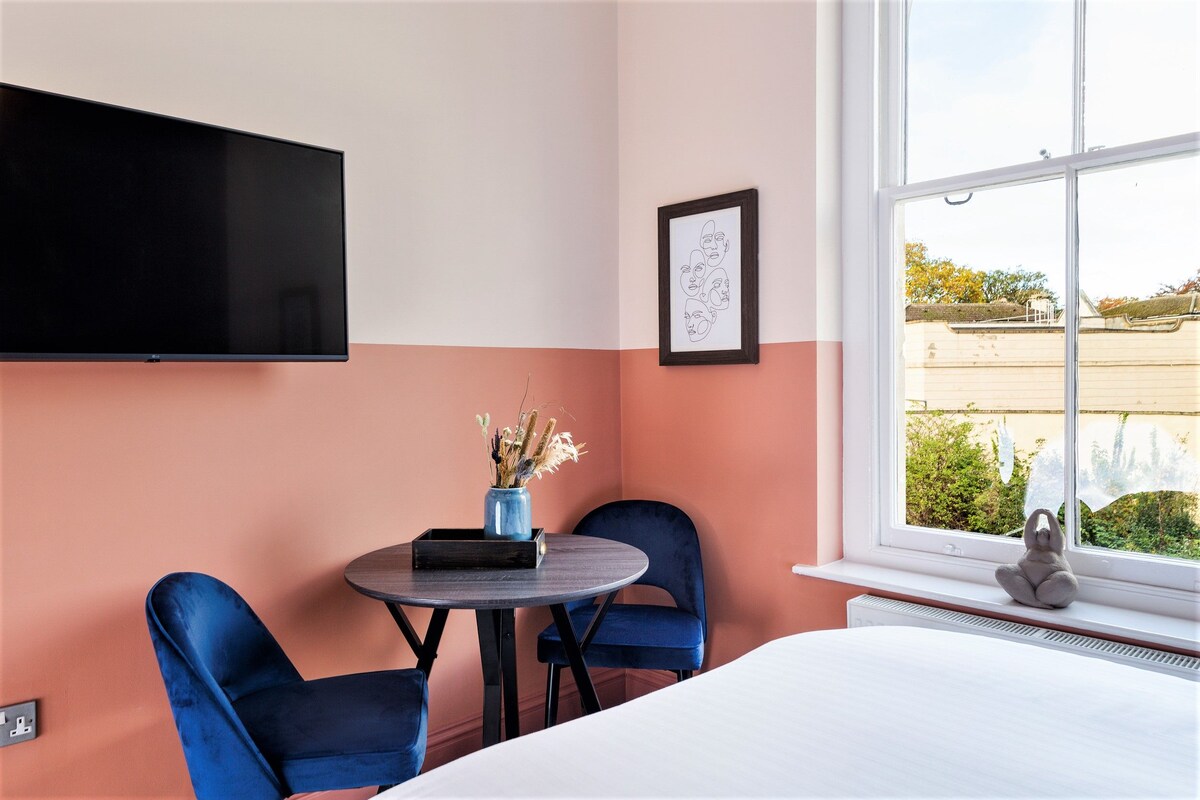 Une chambre d’hôtel avec un lit, un téléviseur fixé au mur, une petite table sur laquelle est posé un vase de fleurs, deux chaises bleues et une fenêtre donnant sur l’extérieur.
