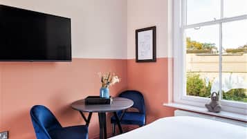 A hotel room with a bed, a TV mounted on the wall, a small table with a vase of flowers, two blue chairs, and a window with a view of the outside.