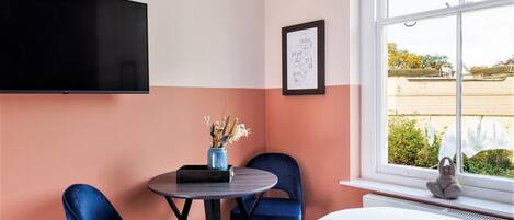 Una habitación de hotel con una cama, un televisor montado en la pared, una mesita con un jarrón de flores, dos sillas azules y una ventana con vistas al exterior.