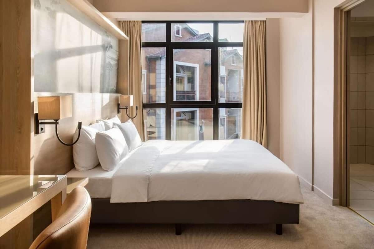 Ein Hotelzimmer mit einem großen Bett, einem Nachttisch, einer Lampe und Blick auf die Stadt durch ein großes Fenster.