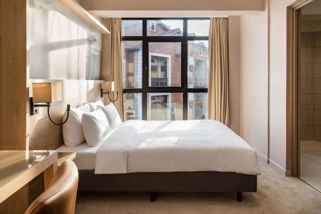 Ein Hotelzimmer mit einem großen Bett, einem Nachttisch, einer Lampe und Blick auf die Stadt durch ein großes Fenster.