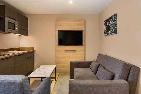 A compact living space with a kitchenette, a flat-screen TV mounted on a wooden panel, a gray sofa, and a small table. - Radisson Serviced Apartments City Center (Antananarivo)