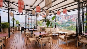 Un área de estar al aire libre con mobiliario de mimbre, lámparas colgantes rojas y vistas a la ciudad.