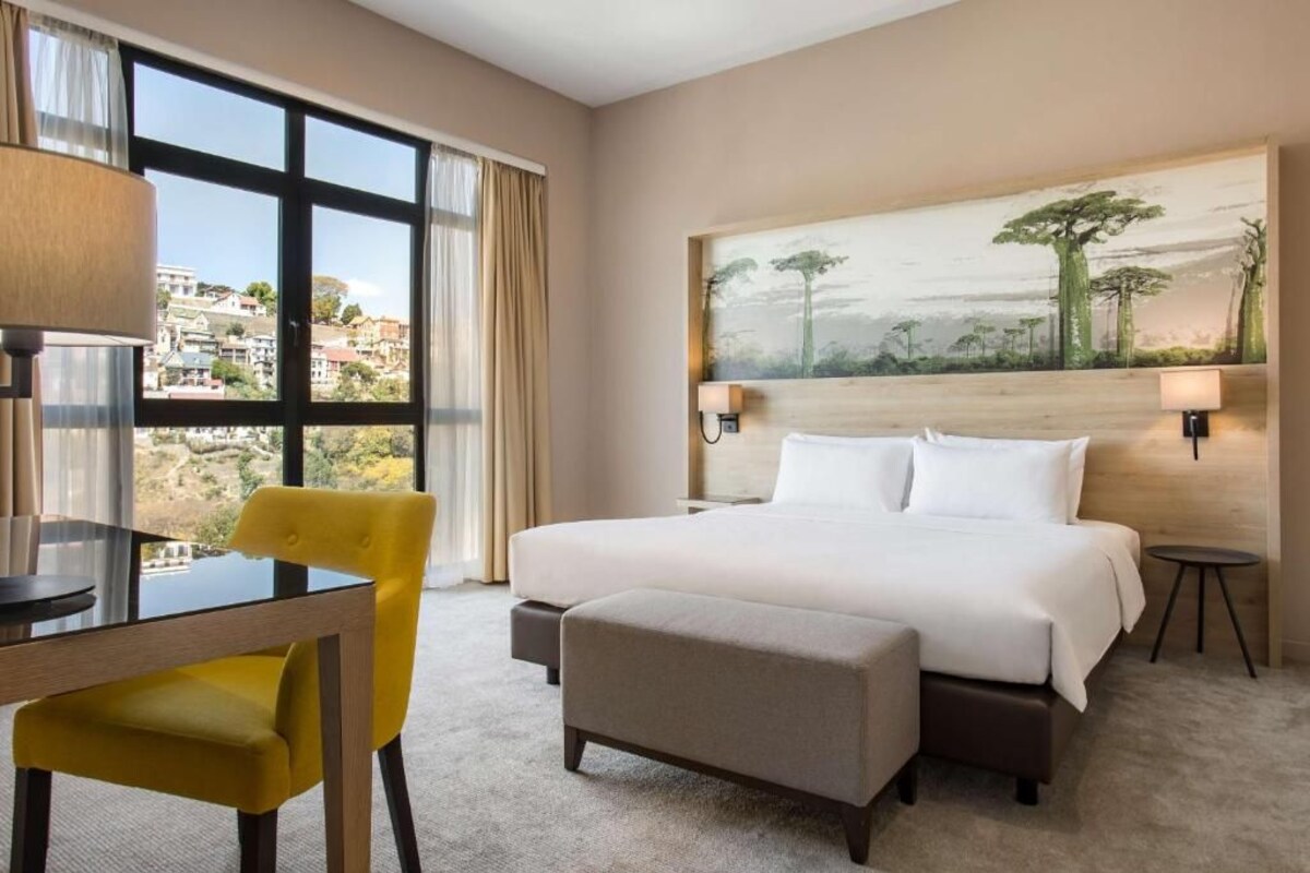 Een hotelkamer met een groot bed, een bureau met stoel, een schilderij aan de muur en uitzicht op de stad door het raam.