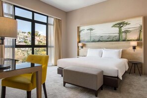 Een hotelkamer met een groot bed, een bureau met stoel, een schilderij aan de muur en uitzicht op de stad door het raam.