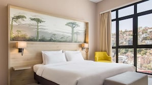 Habitación de hotel con una cama grande, un sofá pequeño, un cuadro en la pared y vistas a edificios a través de un ventanal amplio.