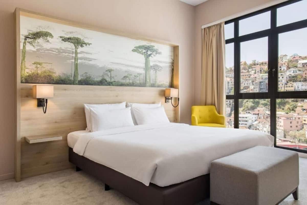 Ein Hotelzimmer mit einem großen Bett, einer kleinen Couch, einem Bild an der Wand und Blick auf Gebäude durch ein großes Fenster.