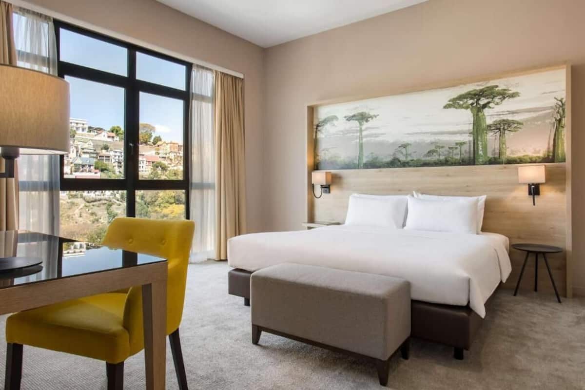 Ein Hotelzimmer mit einem großen Bett, einem Schreibtisch mit Stuhl, einem Bild an der Wand und Blick auf die Stadt durch das Fenster.