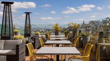 Terraza al aire libre con mesas y sillas, plantas en macetas y vistas a la ciudad.