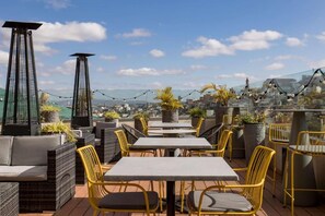 Een terras buiten met tafels en stoelen, planten in potten en uitzicht over de stad.