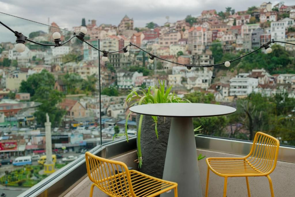Un balcón con dos sillas amarillas, una mesa redonda y luces colgantes, con vistas a la ciudad.