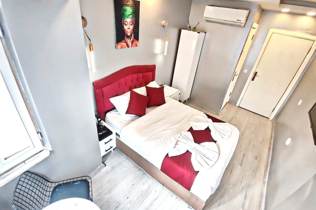 Quarto com cabeceira vermelha, cama com roupa de cama branca e vermelha, uma pequena mesa de cabeceira, ar-condicionado fixado na parede e uma porta.
