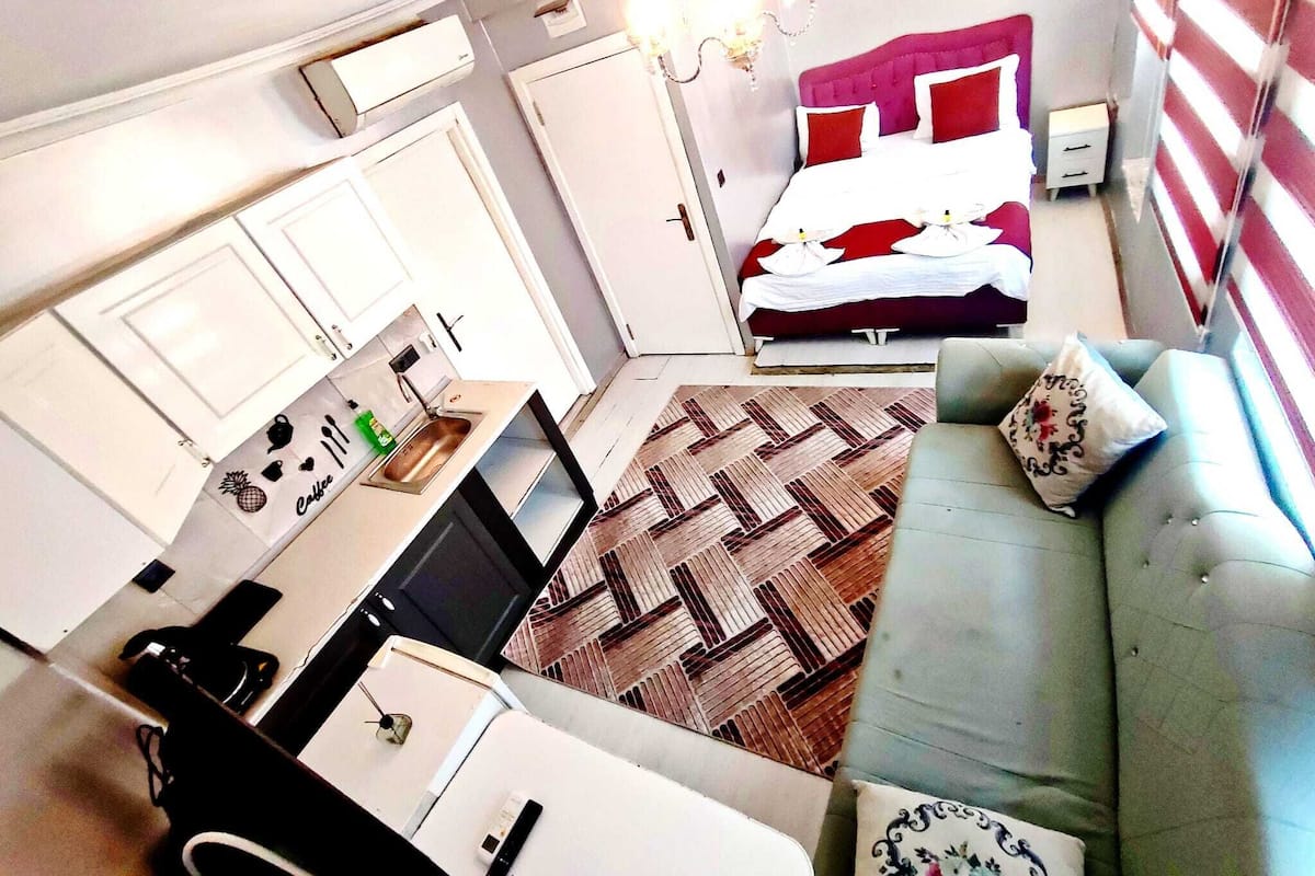 A compact hotel room with a kitchenette, a bed with red and white bedding, a grey sofa, and a patterned rug.