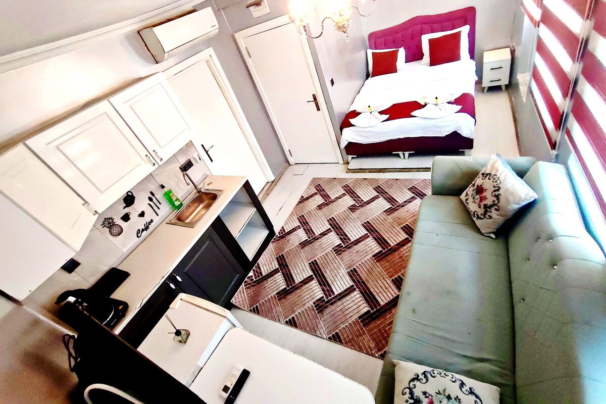 Quarto de hotel compacto com cozinha compacta, cama com roupa de cama vermelha e branca, sofá cinza e tapete estampado.