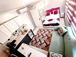 A compact hotel room with a kitchenette, a bed with red and white bedding, a grey sofa, and a patterned rug. - Taksim Capital Hotel (Istanbul)
