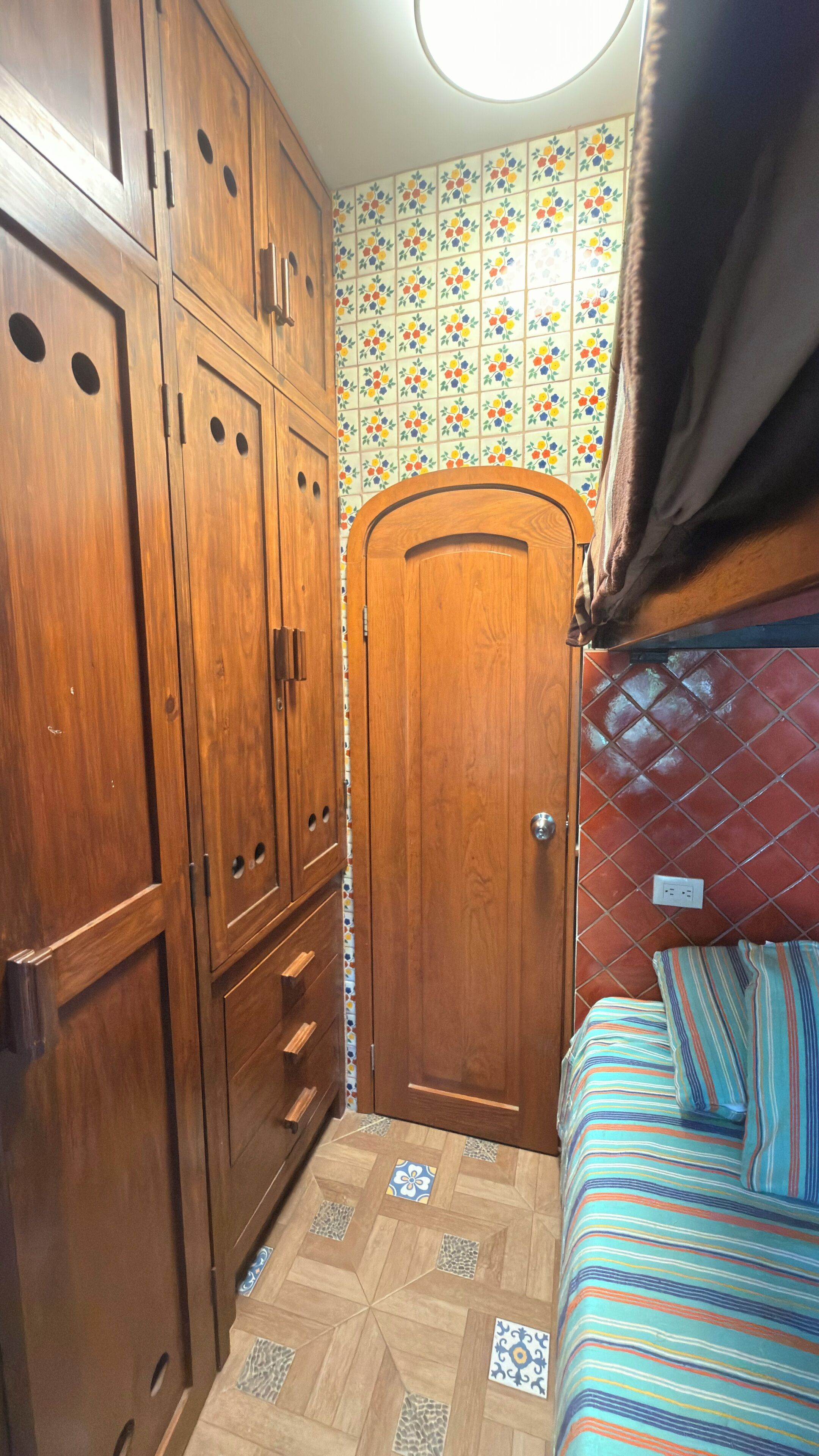 Quarto pequeno com guarda-roupa de madeira, uma porta, cama com roupa de cama listrada e parede revestida de azulejos com padrão floral.