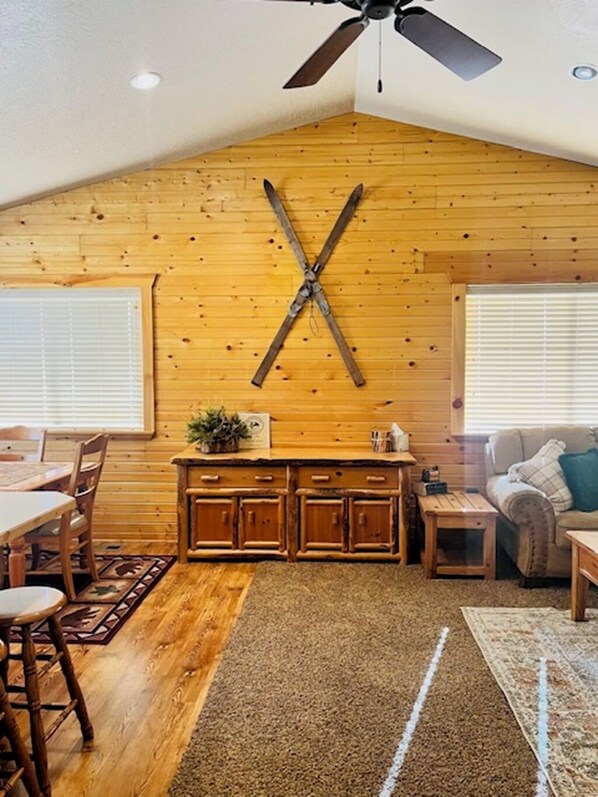 A cozy cabin interior with wooden walls, a dining area, a kitchen with wooden cabinets, and a ceiling fan.