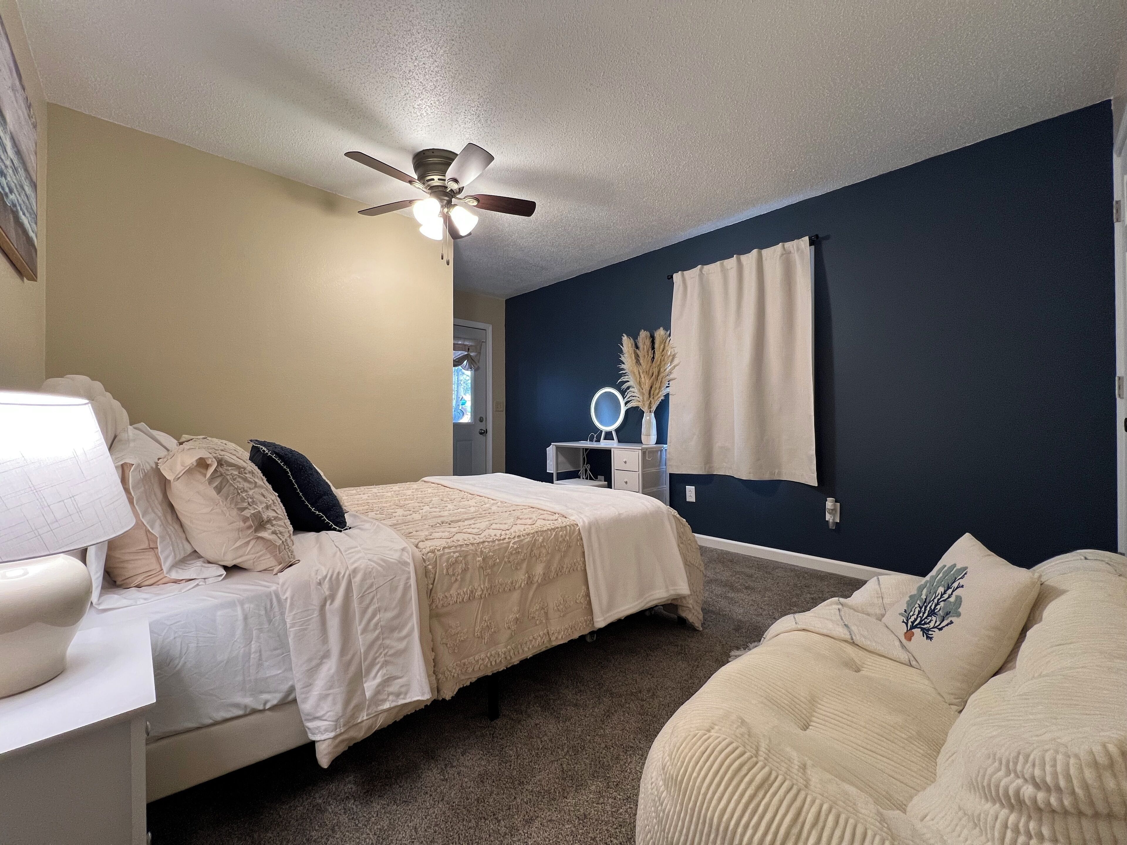 A bedroom with a bed, bedside table, lamp, ceiling fan, dresser, mirror, and a window with curtains.