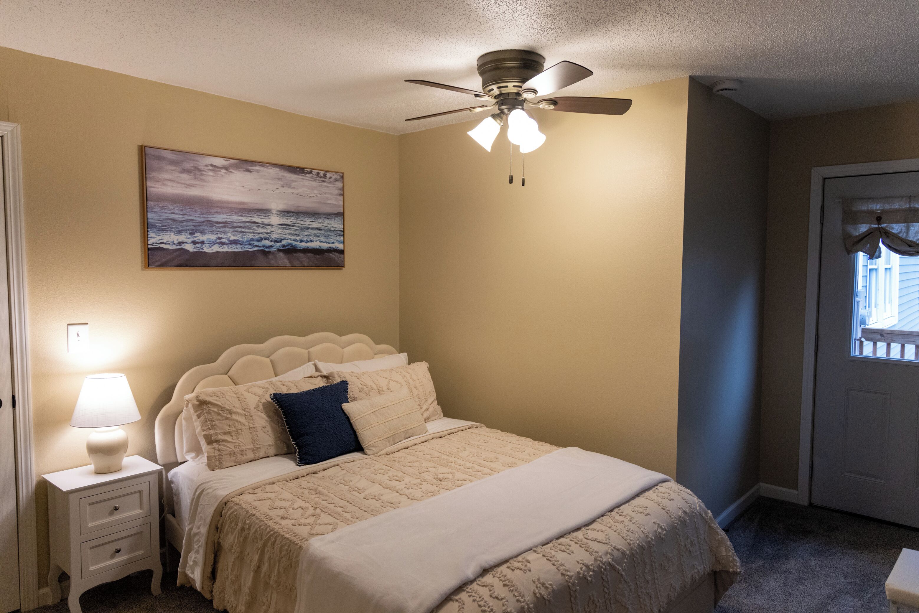 A bedroom with a bed, a ceiling fan, a nightstand with a lamp, and a painting on the wall.