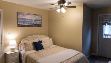 A bedroom with a bed, a ceiling fan, a nightstand with a lamp, and a painting on the wall.