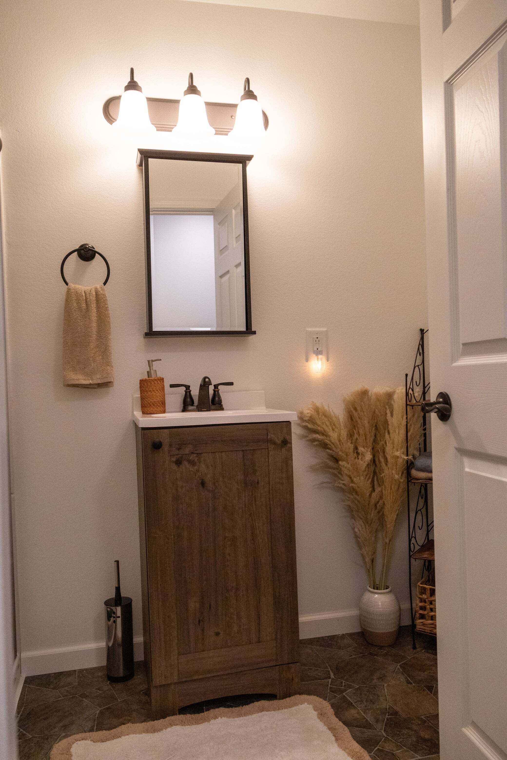 A bathroom with a wooden vanity, a mirror, a towel rack, a vase with pampas grass, and a light fixture.