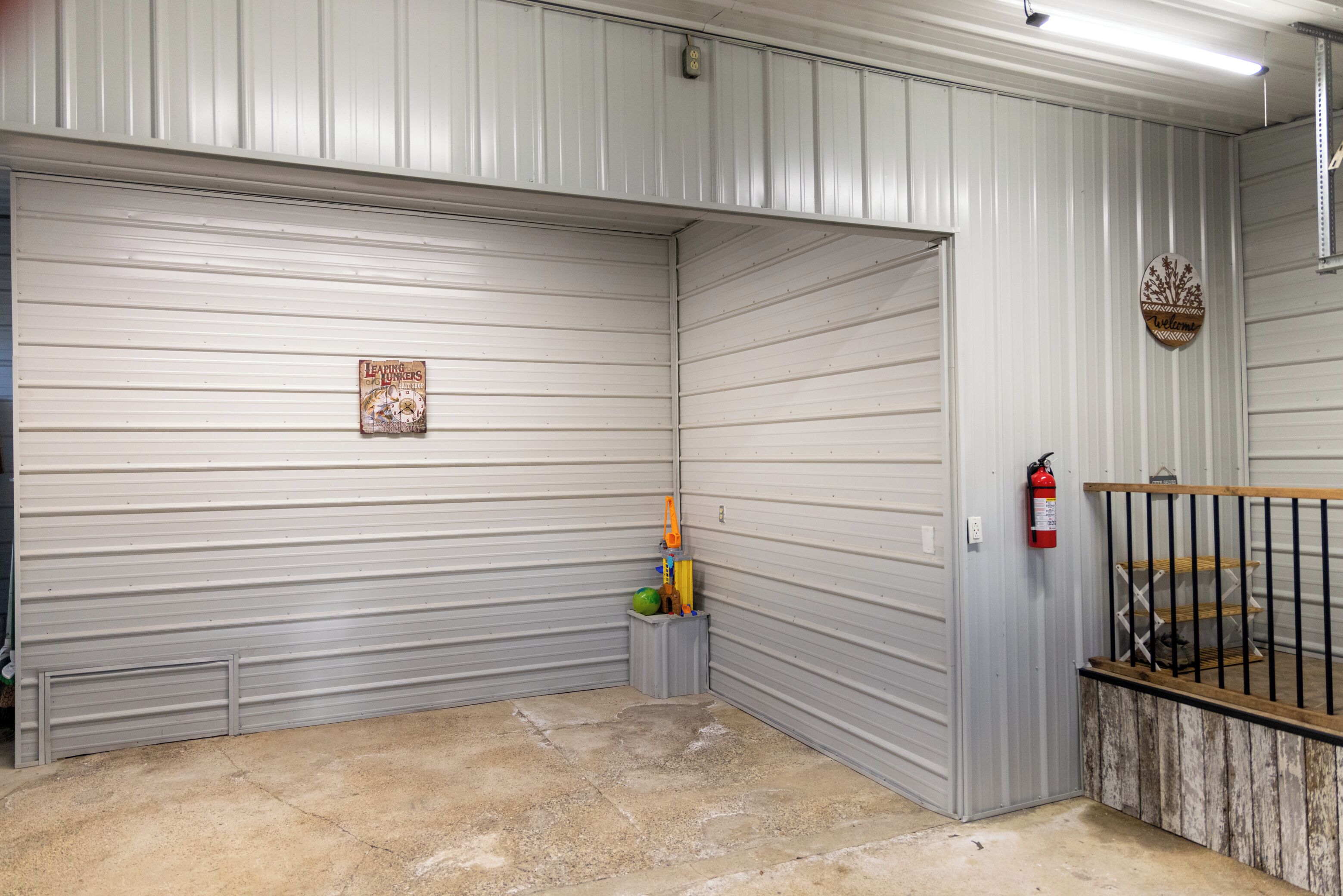 A garage with a closed metal door, a fire extinguisher mounted on the wall, and a small trash bin with a watermelon.