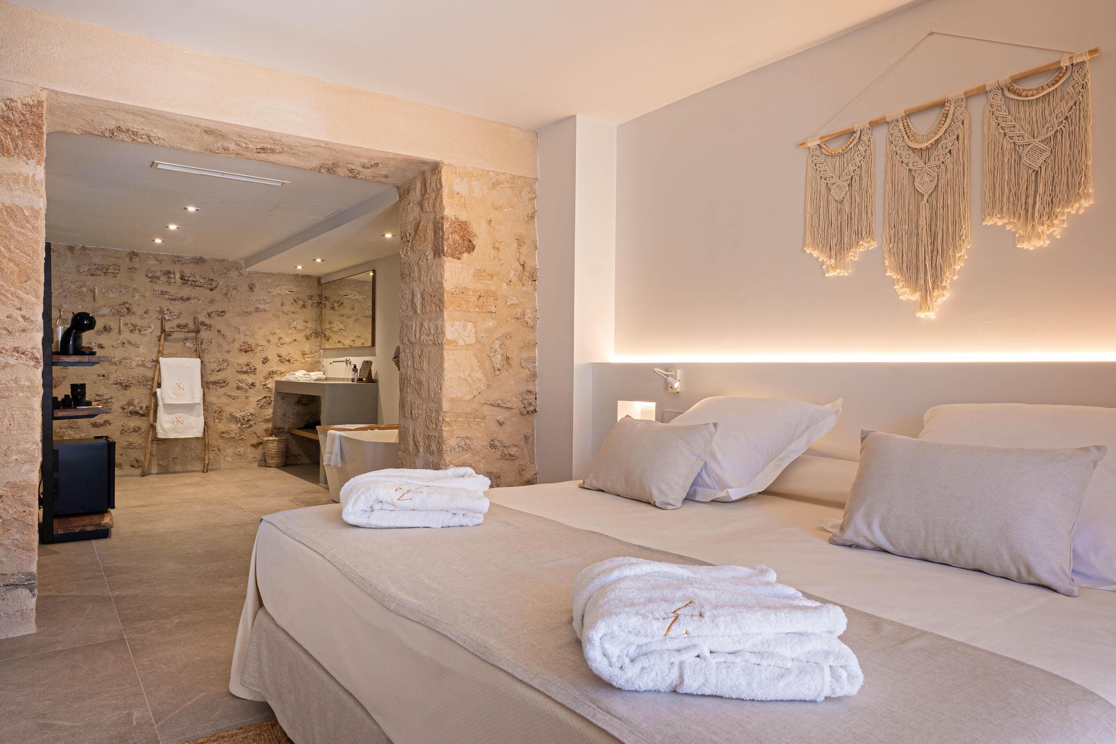 Una habitación de hotel moderna con pared de piedra, una cama grande con ropa blanca, un rincón de descanso con una mesita y una silla, y un baño visible a través de un vano abierto.