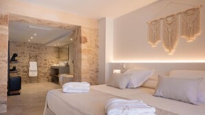 A modern hotel room with a stone wall, a large bed with white linens, a sitting area with a small table and chair, and a bathroom visible through an open doorway. - VILA NOVA SUITES & SPA - DOBLE DELUXE CON BALCÓN (Capdepera)