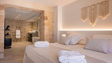 Una habitación de hotel moderna con pared de piedra, una cama grande con ropa blanca, un rincón de descanso con una mesita y una silla, y un baño visible a través de un vano abierto.