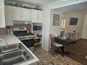 A kitchen with white cabinets, a stove, a microwave, a sink with a bottle rack, and a dining area with a chair and table.